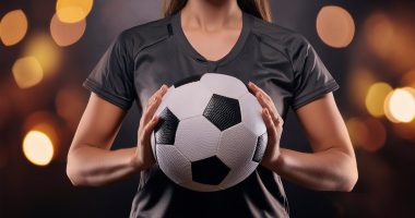 Igualdad futbol femenino. Mujer deportista irreconocible sujeta un balón de fútbol entre sus manos con fondo oscuro y desenfocado de fondo. La atleta lleva una camiseta deportiva negra.